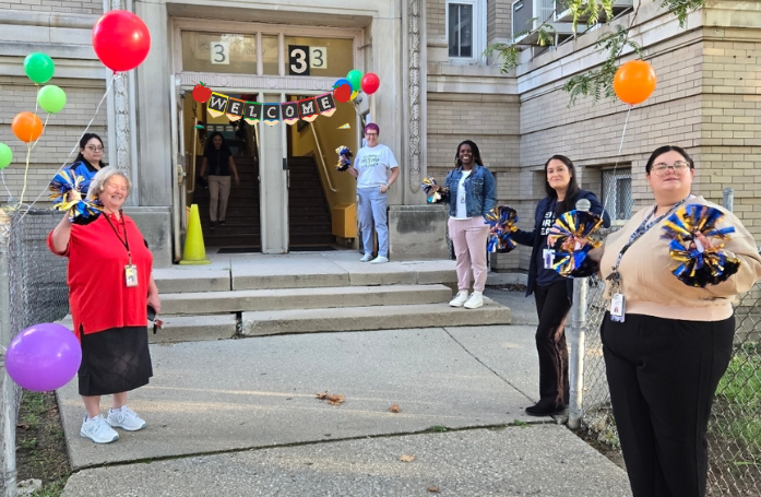  adults lined up along a fence with balloons holding pom poms